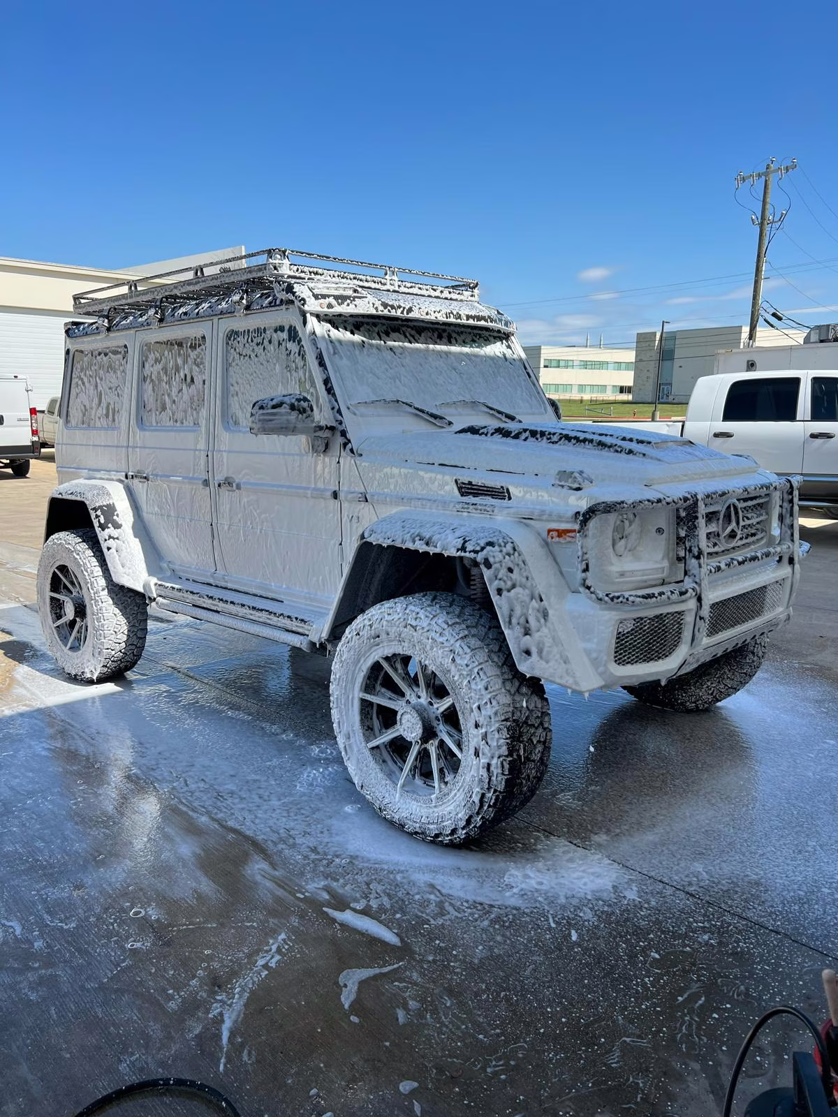 Mercedes-Benz G-Class SUV covered in white foam during a car wash on a sunny day.