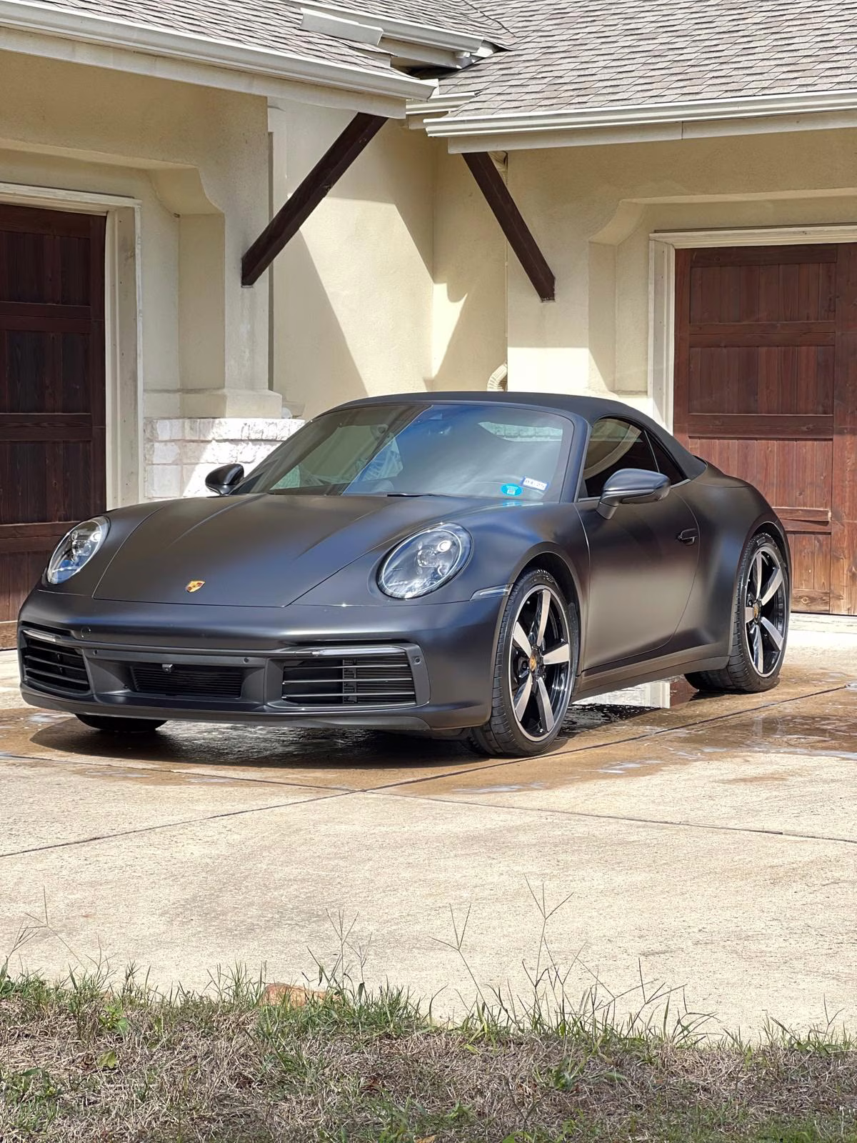 Matte black Porsche convertible parked in a driveway in front of a beige house with wooden garage doors.