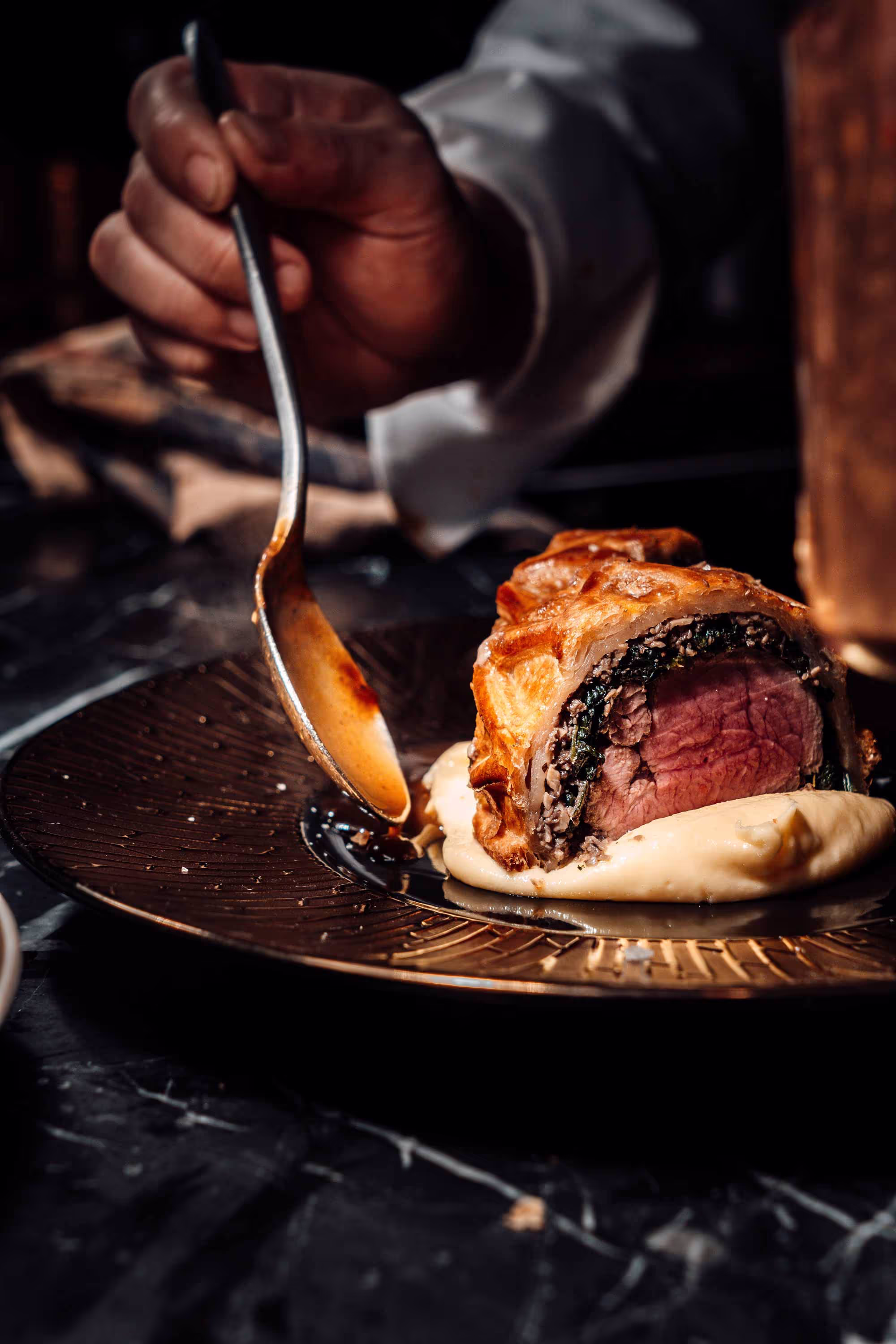 Hand spooning sauce onto plated beef Wellington served on mashed potatoes.