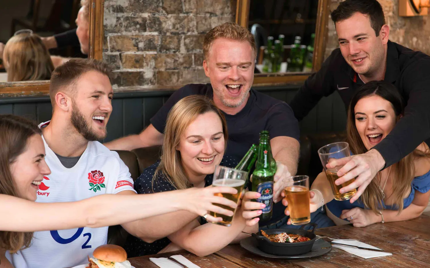 Group of six friends sitting around a wooden table cheer with glasses of beer and bottles in a cozy pub setting.