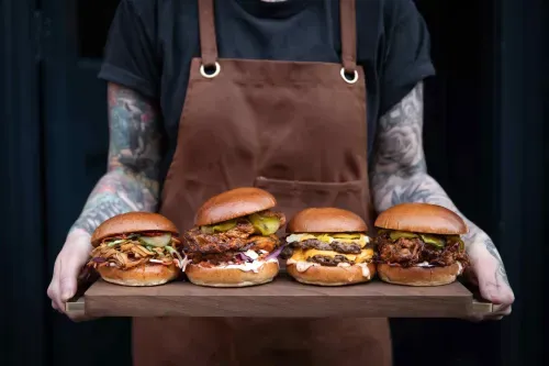 Person with tattooed arms wearing a brown apron holding a wooden tray with four different gourmet burgers.