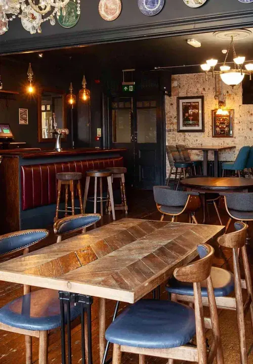 Cozy pub interior with wooden tables, blue cushioned chairs, bar stools, hanging pendant lights, and decorative plates on the wall.