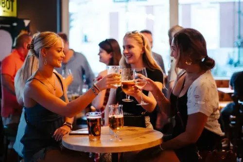 Three women sitting at a wooden table in a lively bar, smiling and clinking glasses filled with beer and wine.