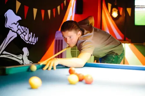 Man aiming with cue stick to hit the cue ball on a pool table in a dimly lit room with skeleton mural and bunting flags.