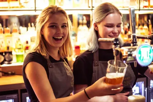 Two female bartenders smiling; one pours beer from a tap into a glass at a bar.