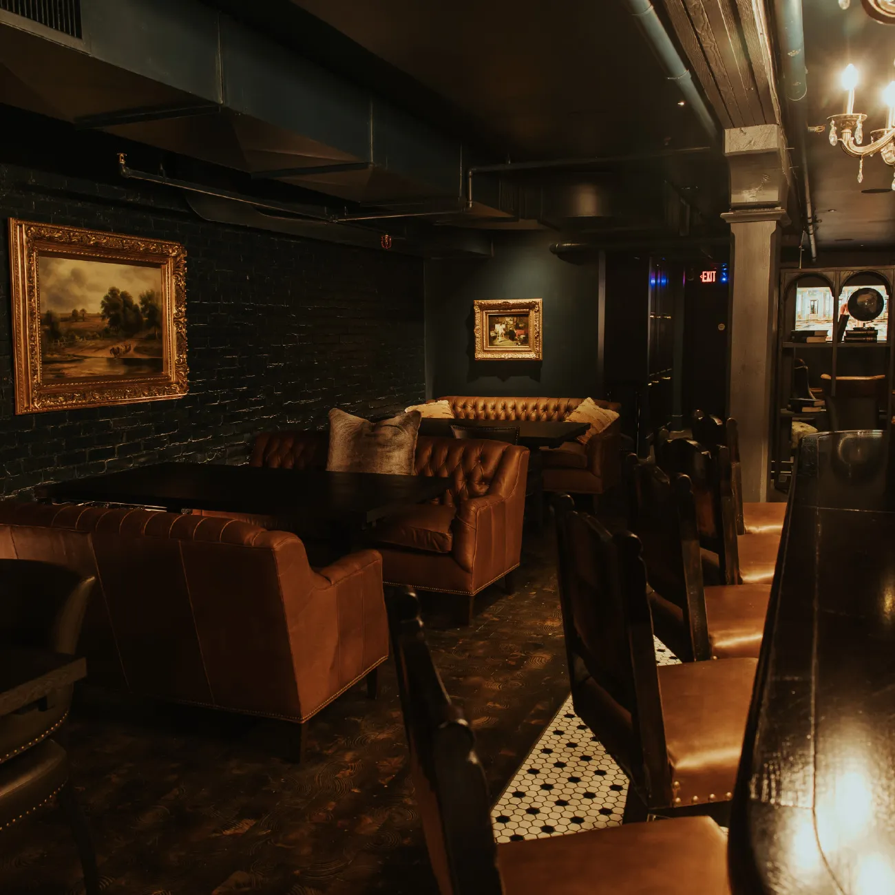 Dimly lit lounge area with brown leather chairs and couches, dark walls, framed paintings, and a black bar with leather stools.