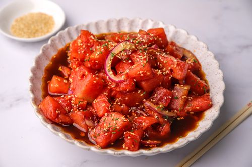 A vibrant bowl of cubed watermelon, thinly sliced red onion, and golden toasted sesame seeds coated in a glossy, sweet and spicy dressing.