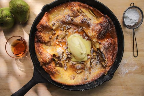 A golden, puffy Dutch baby pancake in a cast-iron pan, topped with spiced pears, walnuts, and a drizzle of maple syrup, ready to serve.