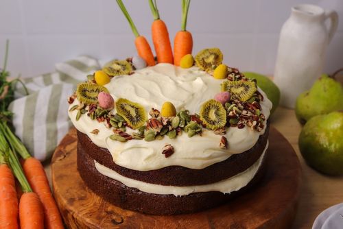 Two-layer carrot cake topped with baby carrots, mini Easter eggs, pumpkin seeds, and dehydrated kiwifruit, on a rustic wooden board.