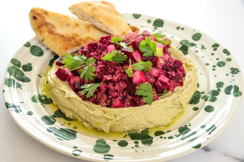 Bowl of creamy avocado hummus topped with paprika, surrounded by beetroot and apple tabbouleh with a drizzle of lemon dressing – a fresh, seasonal side dish.