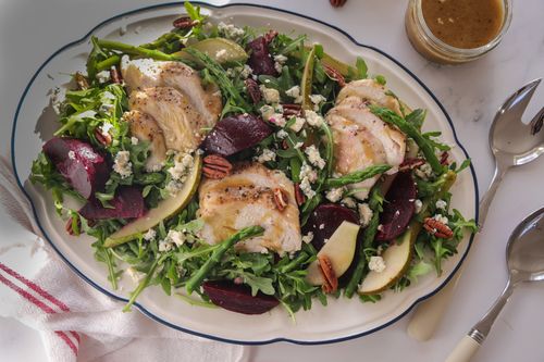 A colourful seasonal salad with sliced chicken breast, roast beetroot wedges, asparagus, pear, pecans and blue cheese on a white plate.