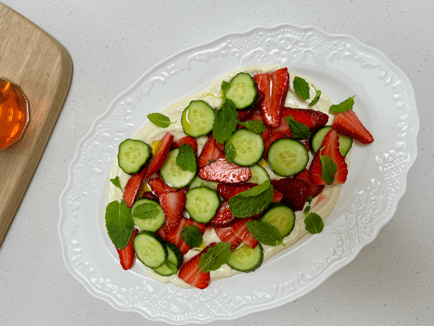 A swirl of whipped feta topped with marinated strawberries, sliced cucumber, and mint leaves, served with crusty bread on a ceramic plate.