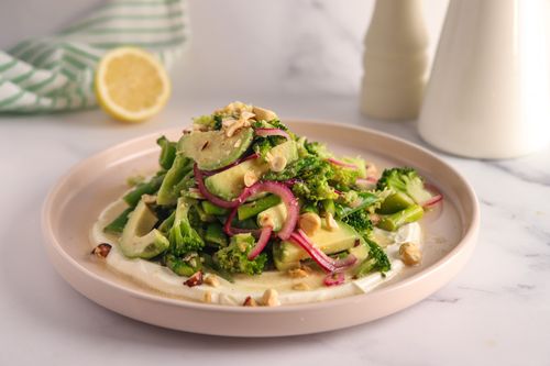 Vibrant green salad with blanched asparagus, broccoli, and snow peas, topped with sliced avocado, pink pickled onions, and chopped toasted hazelnuts, served over a swirl of creamy yoghurt sauce.