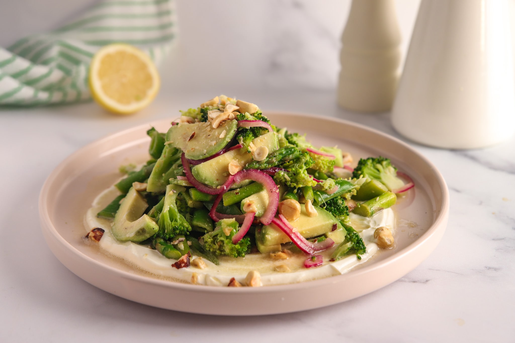 Vibrant green salad with blanched asparagus, broccoli, and snow peas, topped with sliced avocado, pink pickled onions, and chopped toasted hazelnuts, served over a swirl of creamy yoghurt sauce.