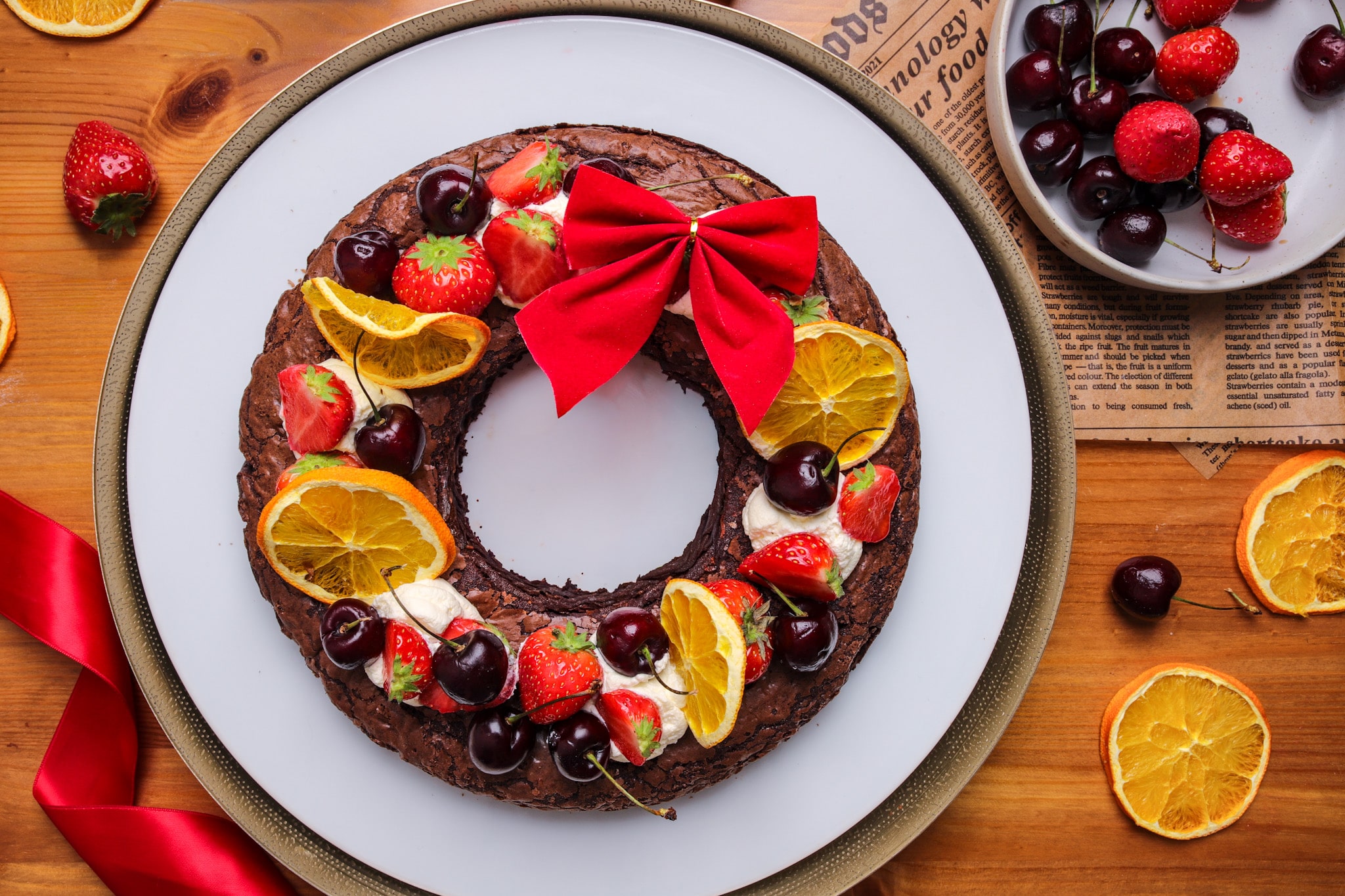 Chocolate brownie wreath topped with whipped cream and fresh summer fruits like cherries, strawberries, and plums, with festive dried orange slices.