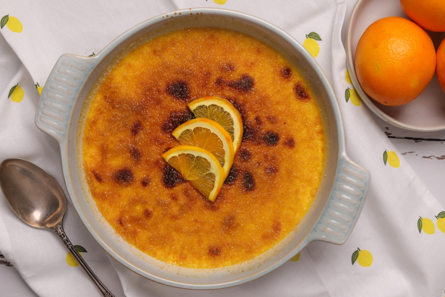 Golden-orange crème brûlée set in a white sharing dish, with a glossy caramelised sugar top and orange zest scattered around.