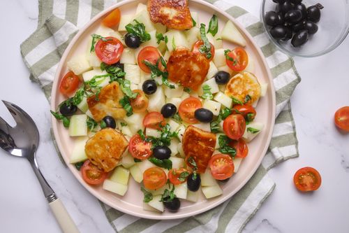 Golden pan-fried halloumi slices glazed with hot honey arranged over cubed melon, cherry tomatoes and black olives, finished with torn fresh basil leaves on a large serving plate