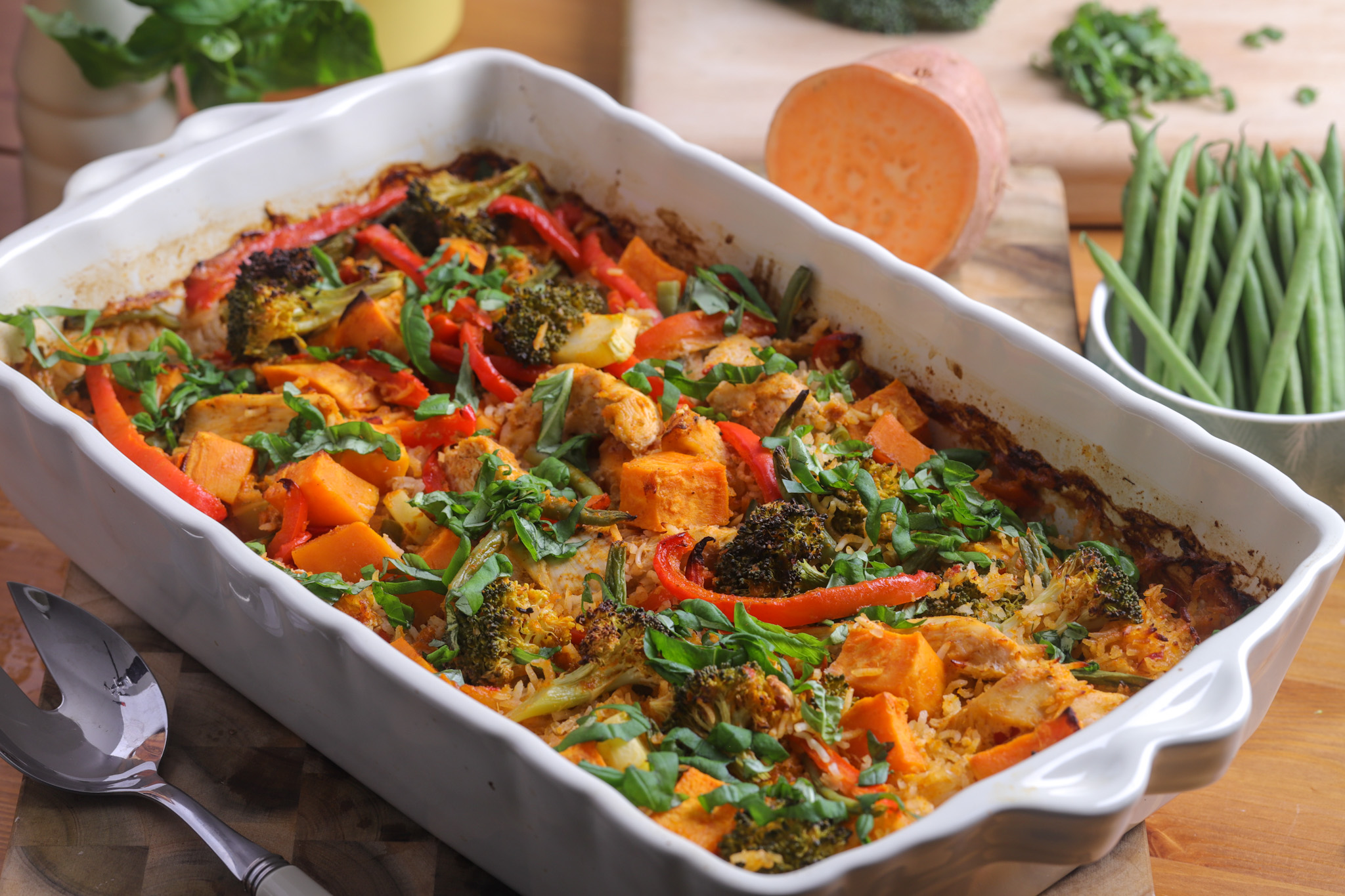A golden Thai-style chicken and vegetable bake straight from the oven, with rice, broccoli, red pepper, and kumara in a coconut milk sauce, topped with fresh basil and a wedge of lime.