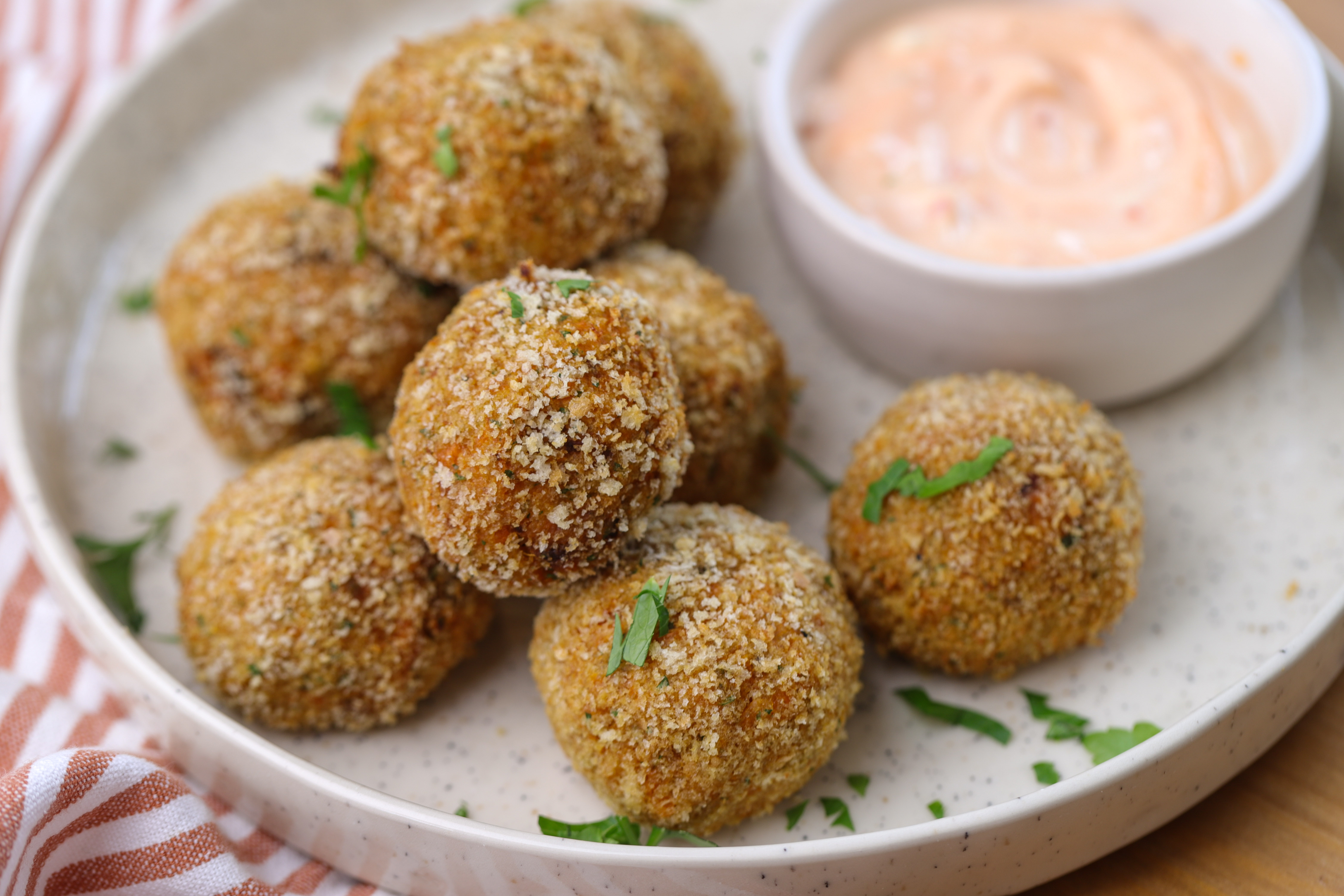 Golden crumbed pumpkin bites stacked on a board with melted mozzarella oozing from the centre, served with a small bowl of spicy mayo.