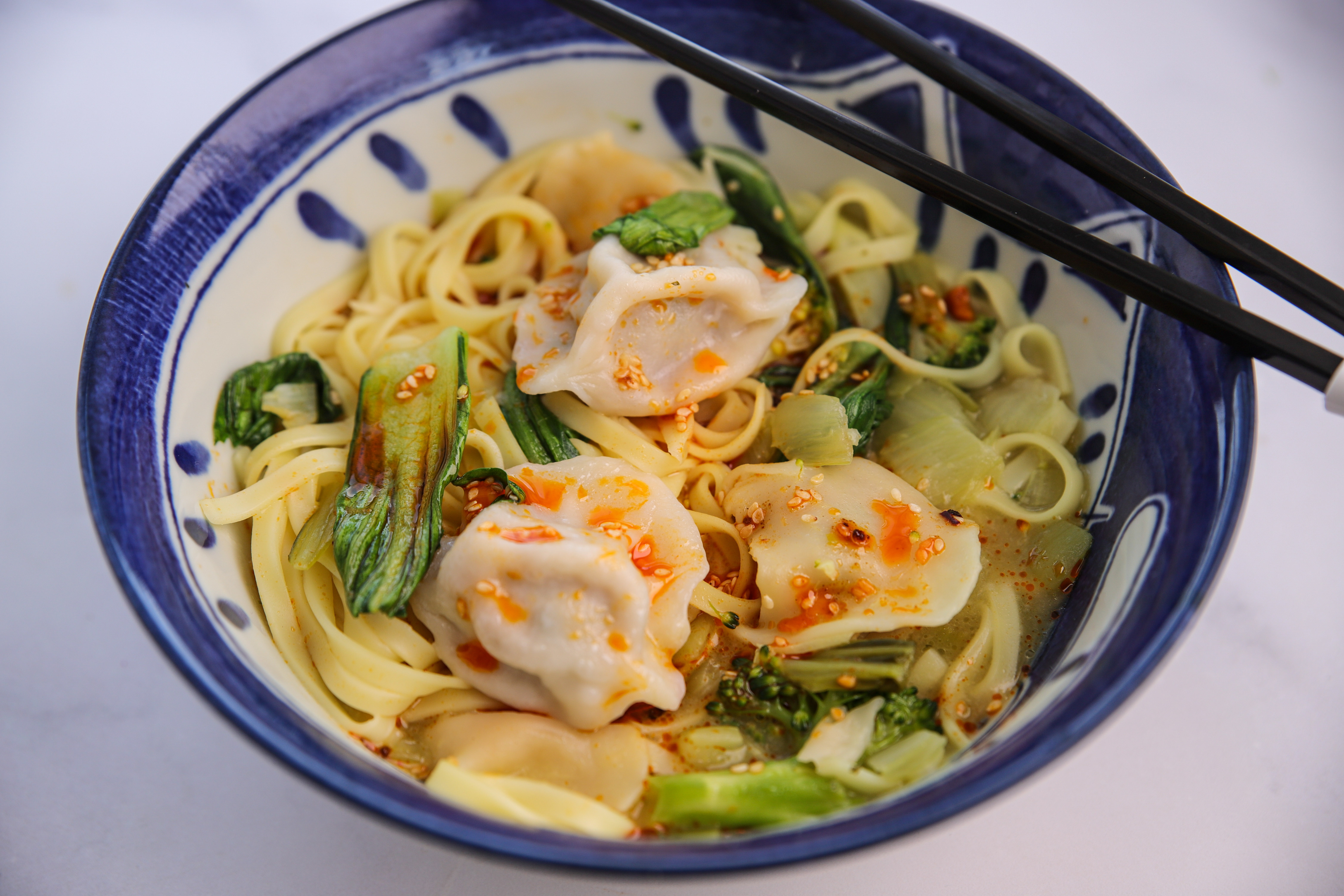  Steaming bowl of dumpling noodle soup with broccoli, pak choi, and egg noodles, drizzled with chilli oil and served from a rice cooker