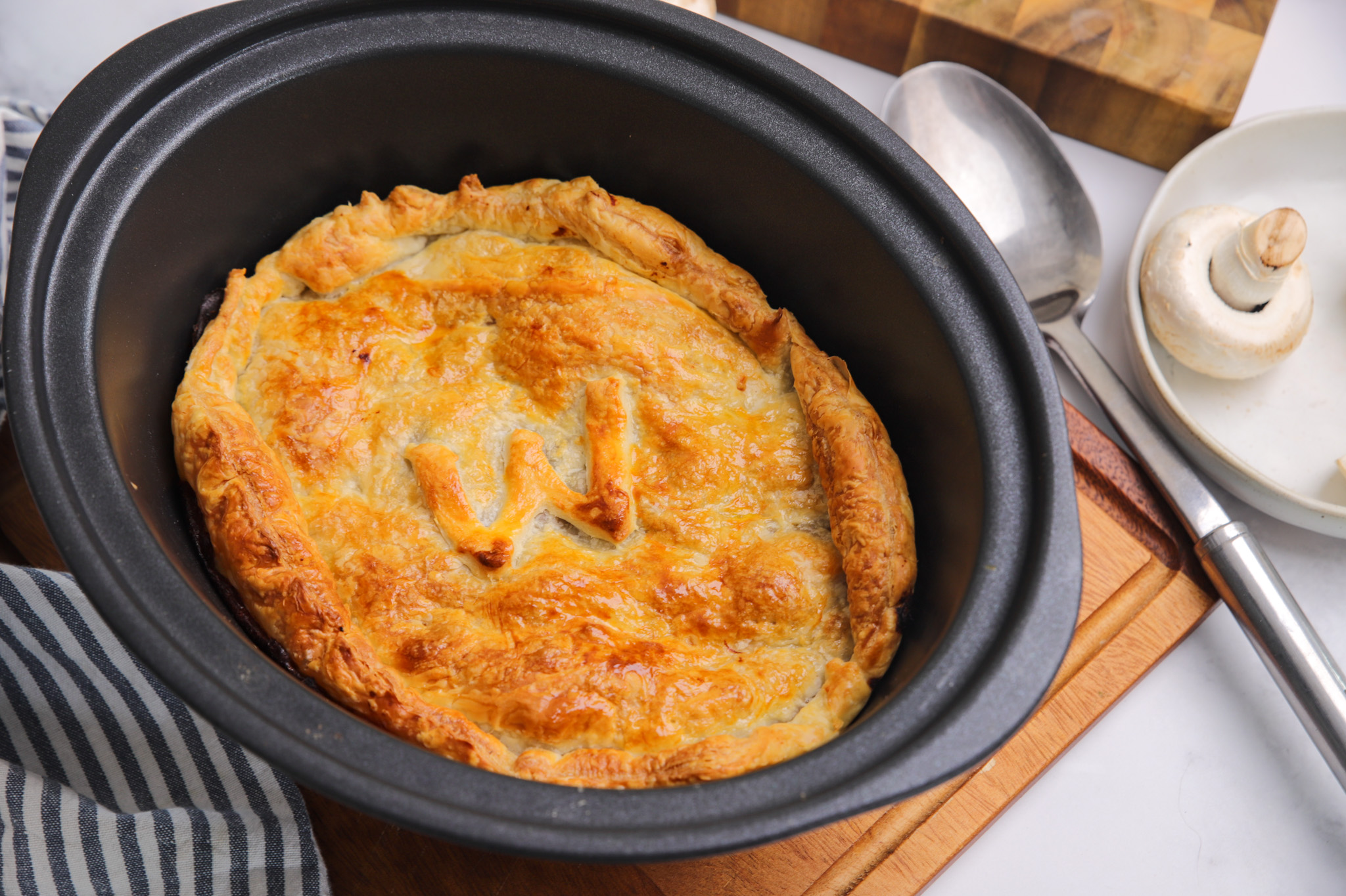 Slow cooker beef pie with golden puff pastry lid, crimped edges and steam holes, served in a rustic ceramic dish with tender beef and vegetable filling visible at the edge