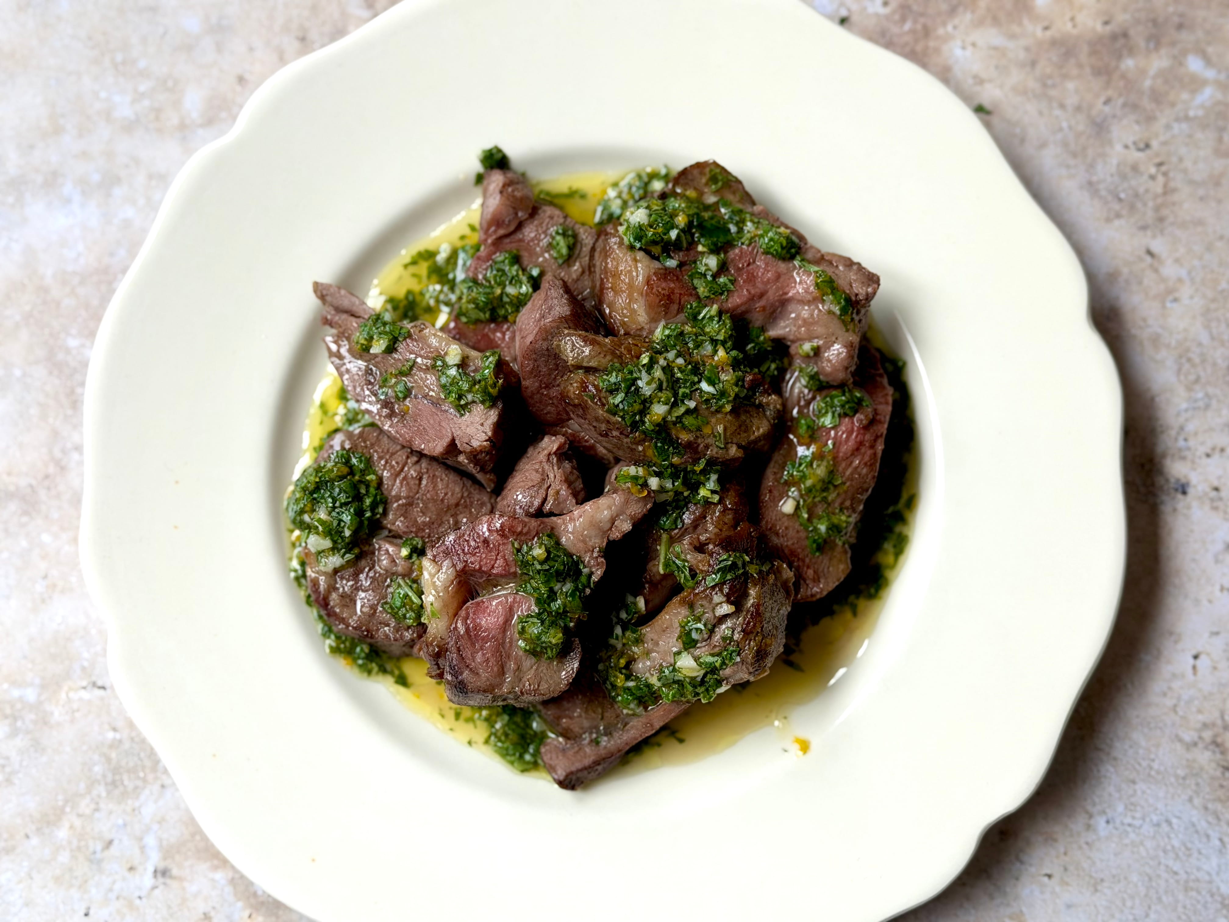 Sliced rolled lamb shoulder fanned across a white platter, spooned with a green mint and basil gremolata flecked with red sumac and lemon zest.