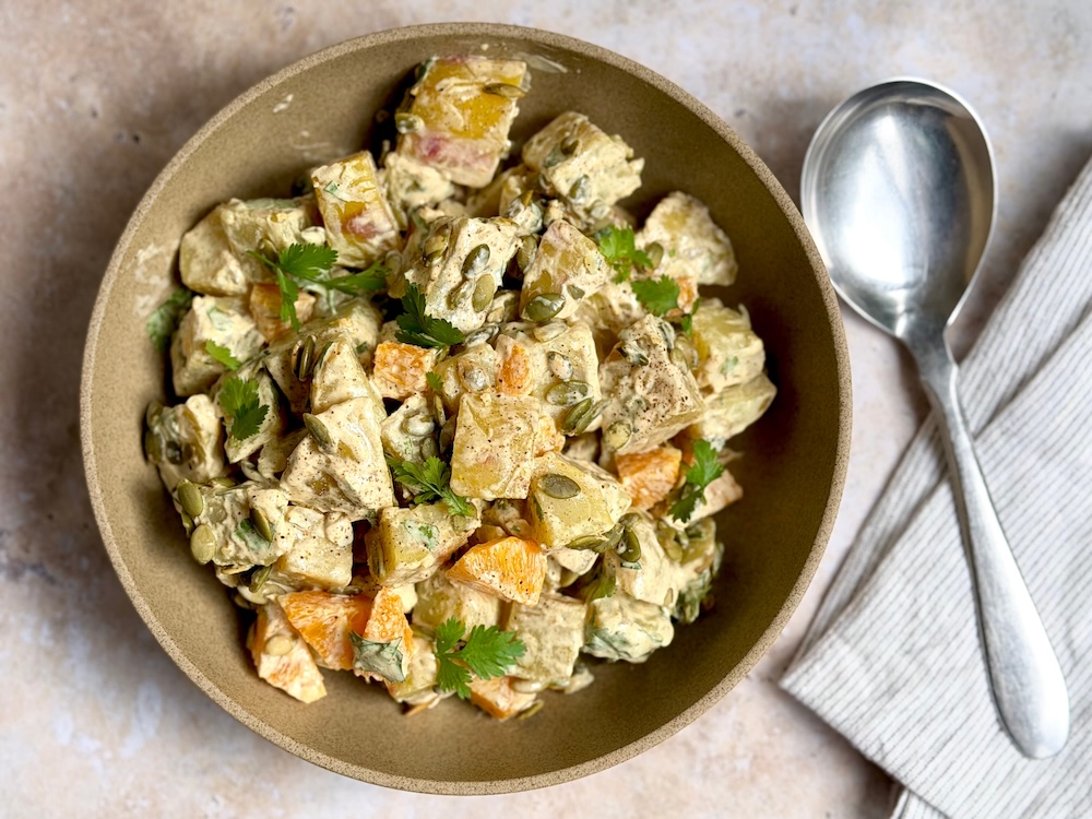 Cubes of boiled kumara tossed in a pale curried sour cream dressing, scattered with orange pieces, toasted pumpkin seeds and fresh coriander in a wide serving bowl.