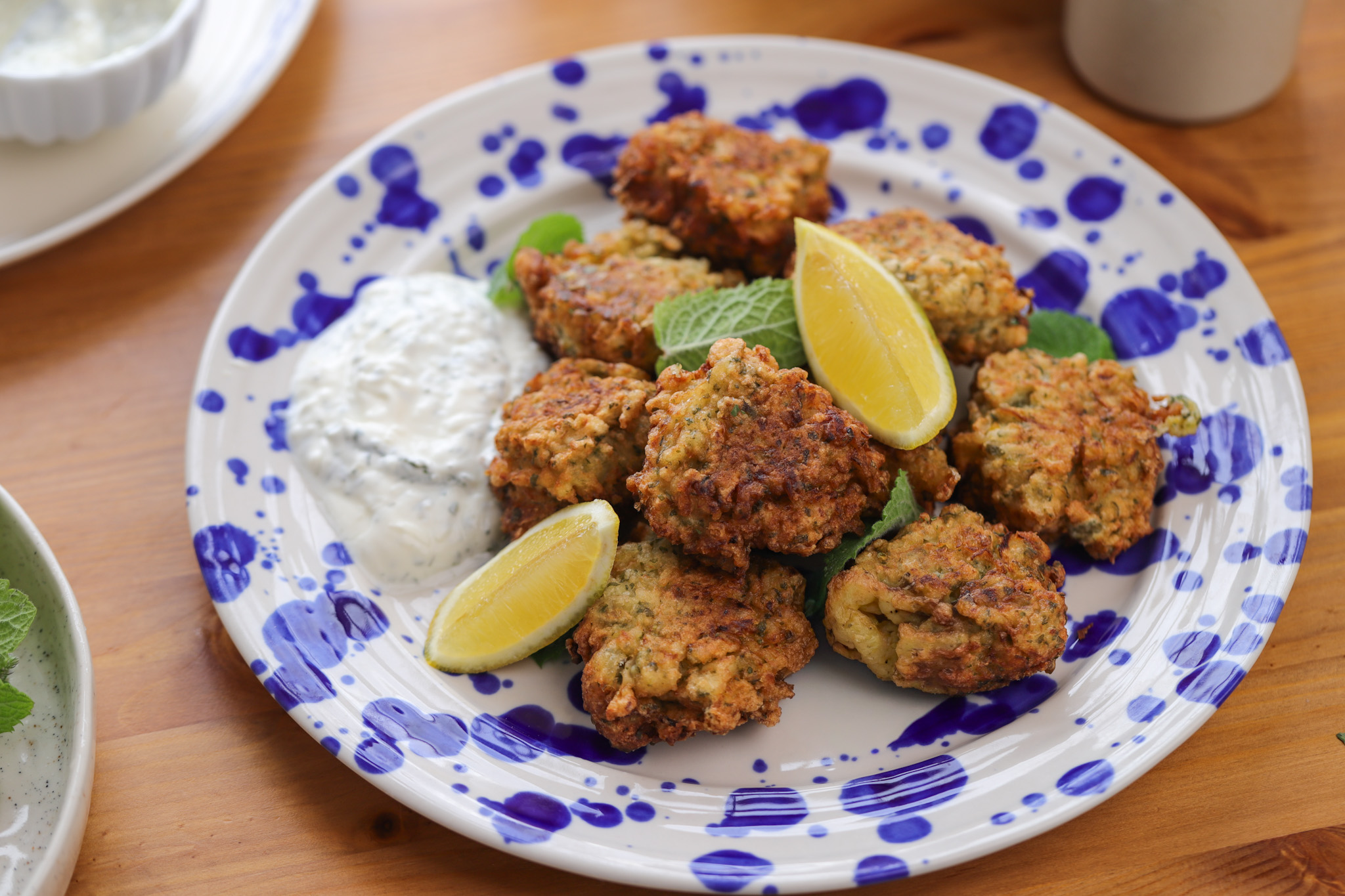 Golden-brown fennel and onion fritters stacked on a plate with a bowl of garlicky yoghurt dip and lemon wedges
