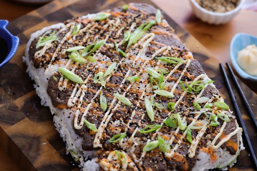 Layered sushi cake with teriyaki beef, avocado and nori, sliced into squares and drizzled with spicy mayo