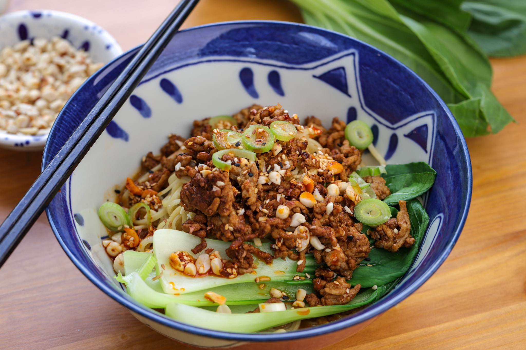Bowl of dan dan noodles topped with crispy pork mince, blanched pak choi, crushed peanuts and sliced spring onion in a glossy tahini and chilli sauce