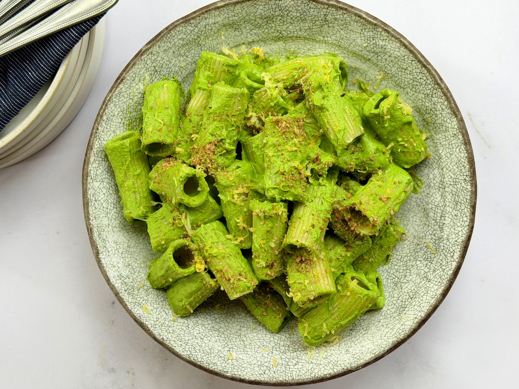 Bowl of rigatoni coated in a smooth green cauliflower and cashew sauce, topped with breadcrumbs and lemon zest.