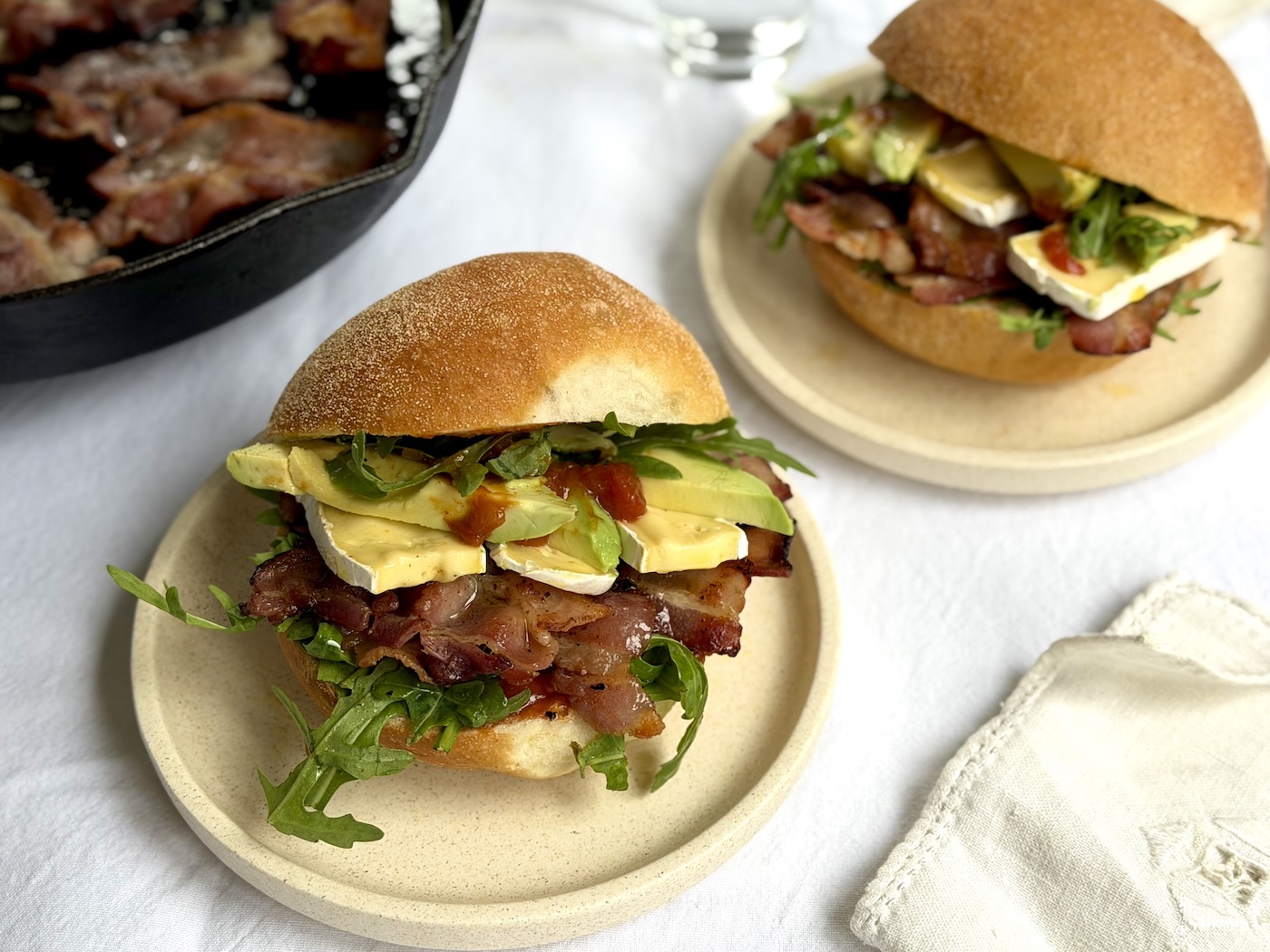 Soft bread bap stacked with crispy bacon, sliced camembert, avocado, rocket and tomato chutney, with more bacon frying in a cast iron skillet behind
