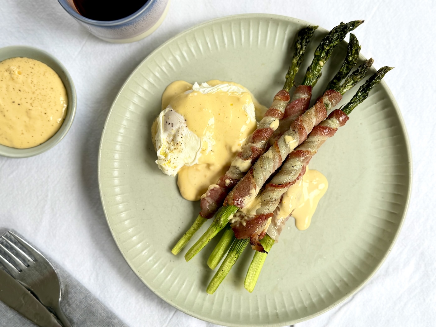 Asparagus spears wrapped in crispy bacon on a sage green plate with a soft poached egg, lemony hollandaise sauce and a side bowl of extra hollandaise