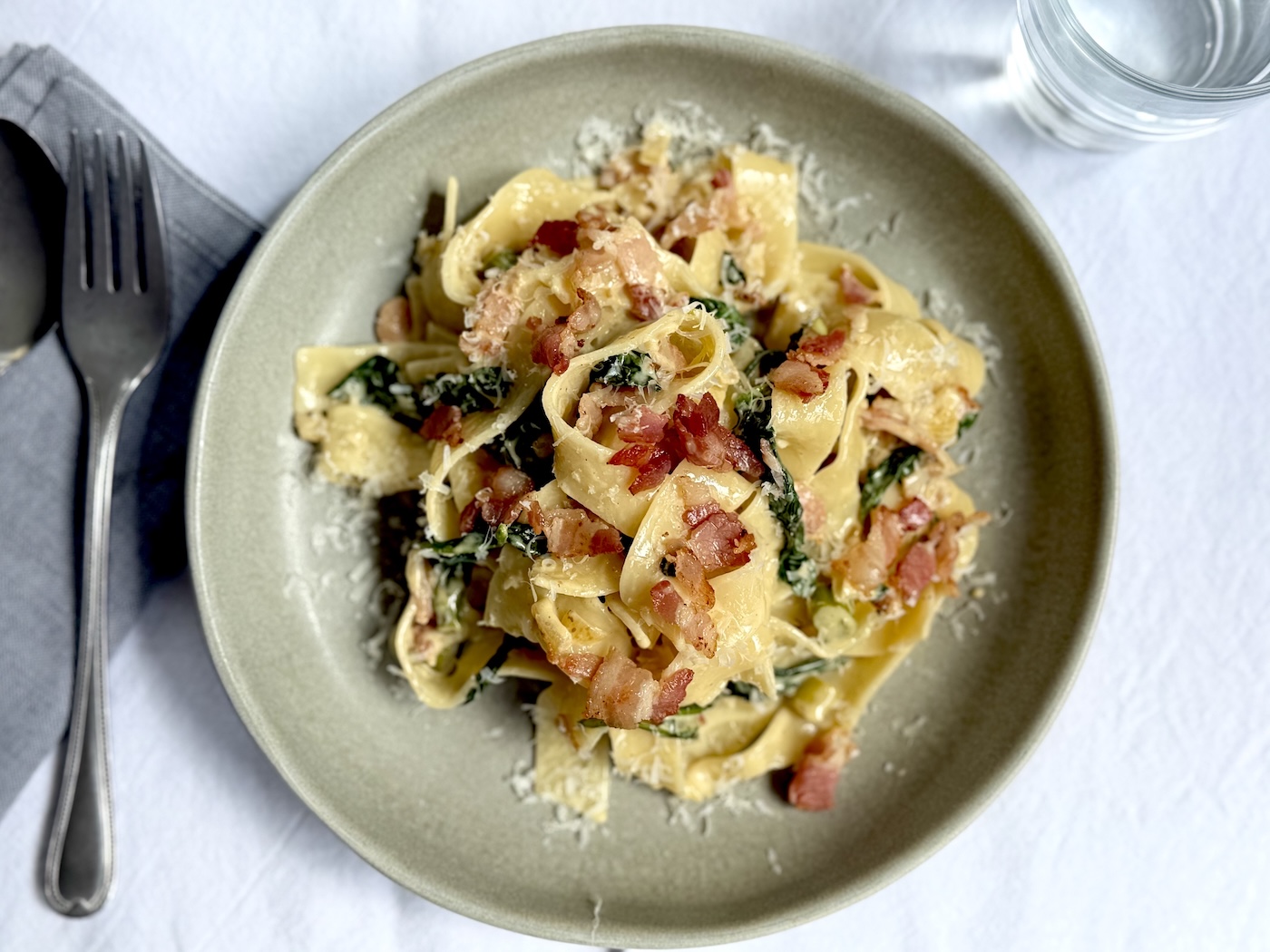 Creamy pappardelle with crispy bacon pieces, wilted baby spinach and asparagus in a grey ceramic bowl, finished with finely grated parmesan