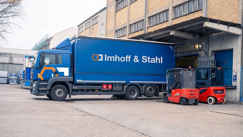 Lkw mit Imhoff & Stahl-Logo beim Beladen an einer Verladerampe, daneben zwei rote Gabelstapler auf dem Firmengelände vor einem Industriegebäude.