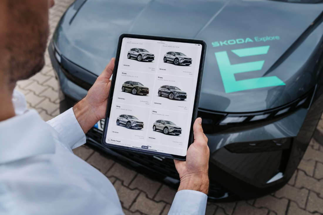 Man holding a tablet displaying a selection of Škoda electric car models in front of a black Škoda car with a green Škoda Explore logo on the hood.