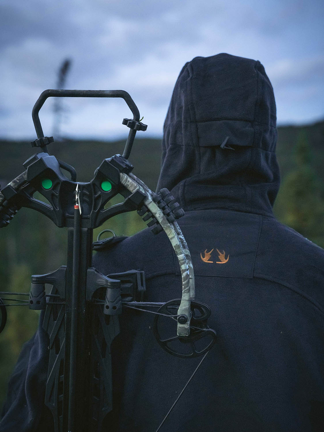 Person wearing a hooded jacket carrying a camouflaged compound bow on their back in an outdoor setting.