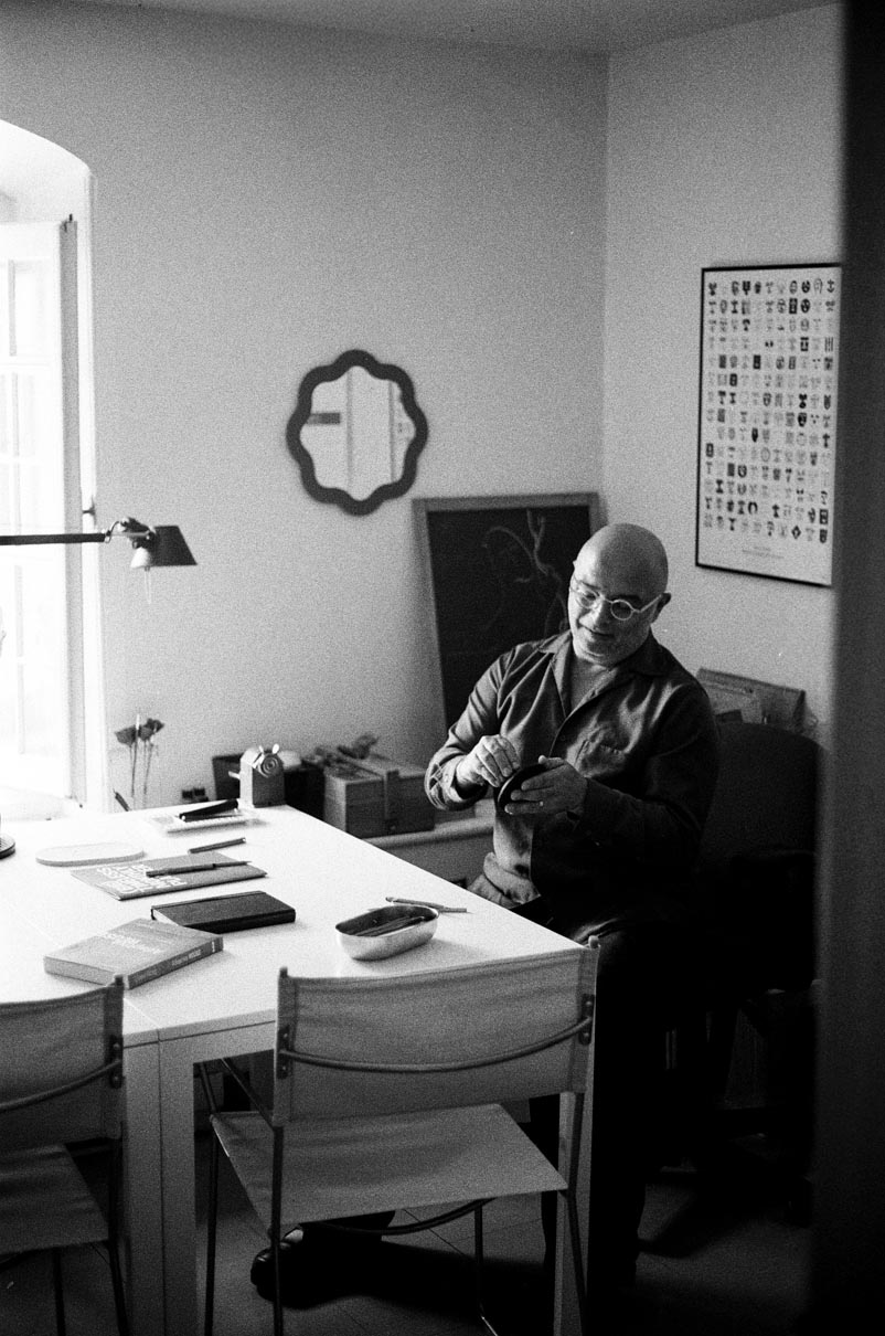 Black and white photo of a bald man with glasses sitting at a table with books and notebooks, inside a room with a window, lamp, and wall decorations.