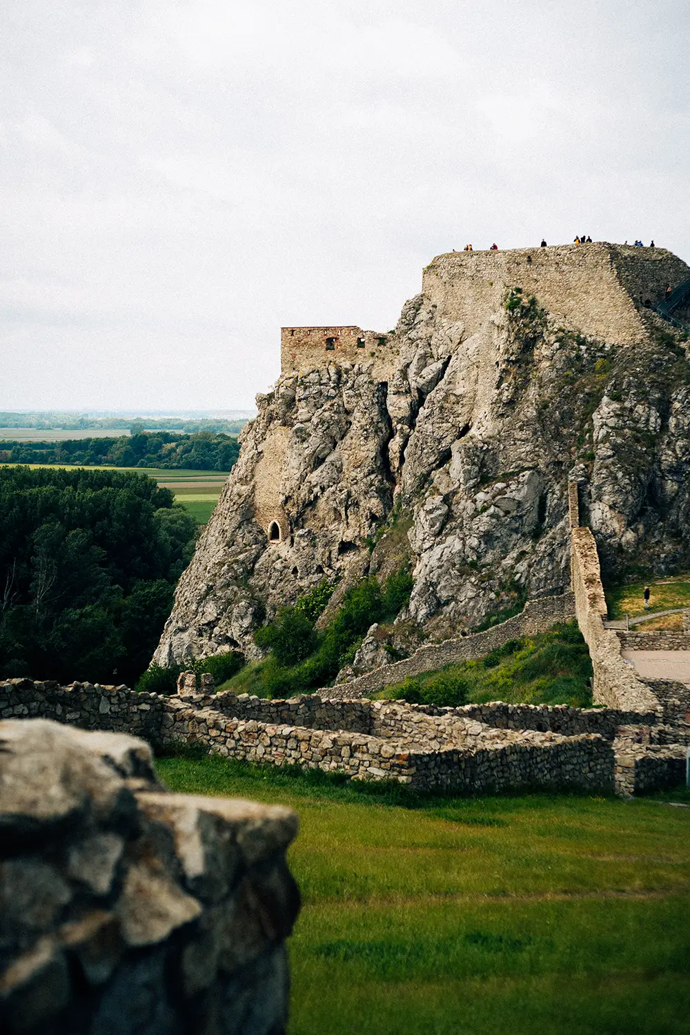 Ancient stone fortress ruins on a rocky hill with green fields and forest in the background.