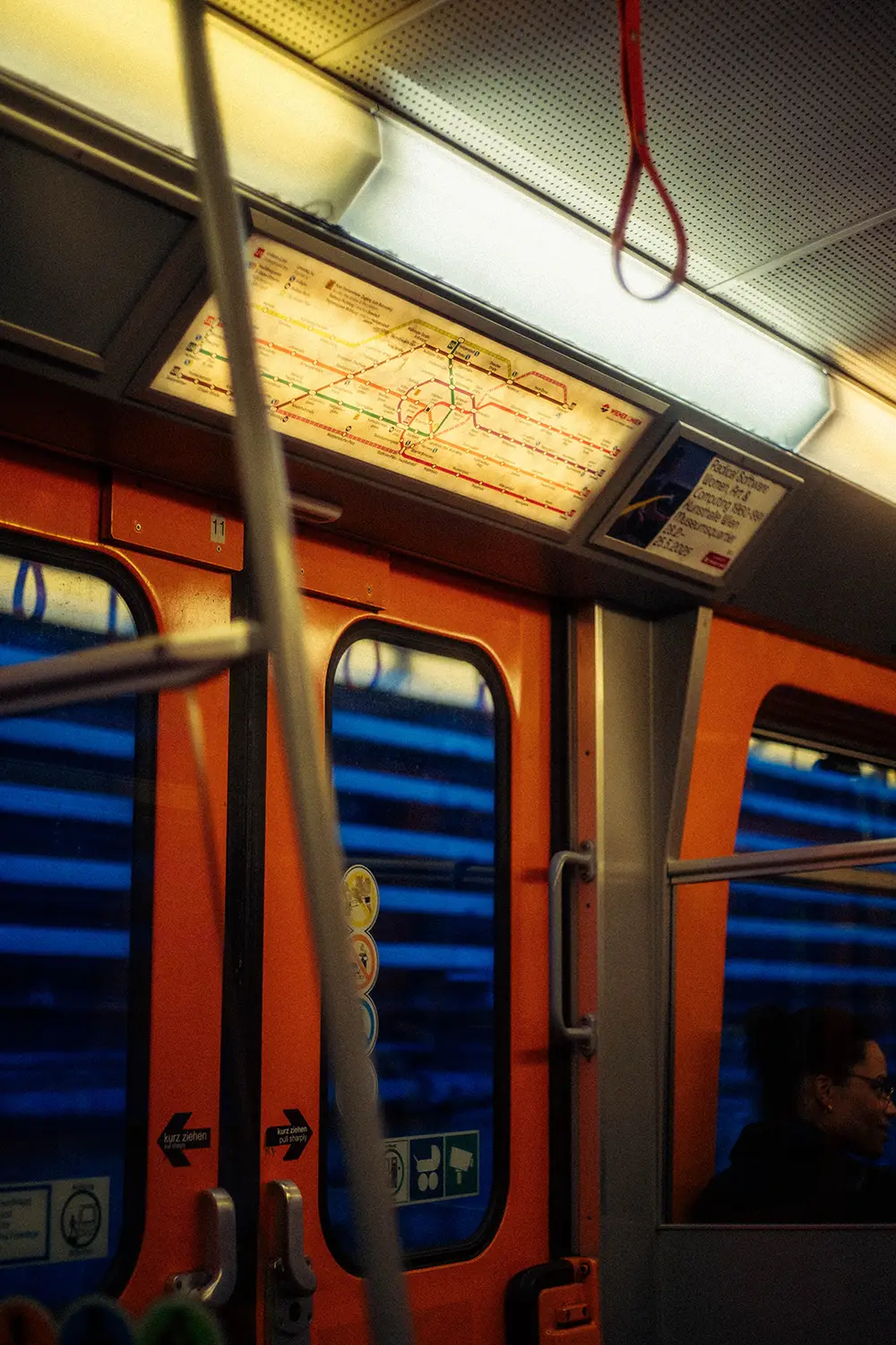 Interior of an orange subway train car with illuminated route map above the doors and a woman sitting by a window.