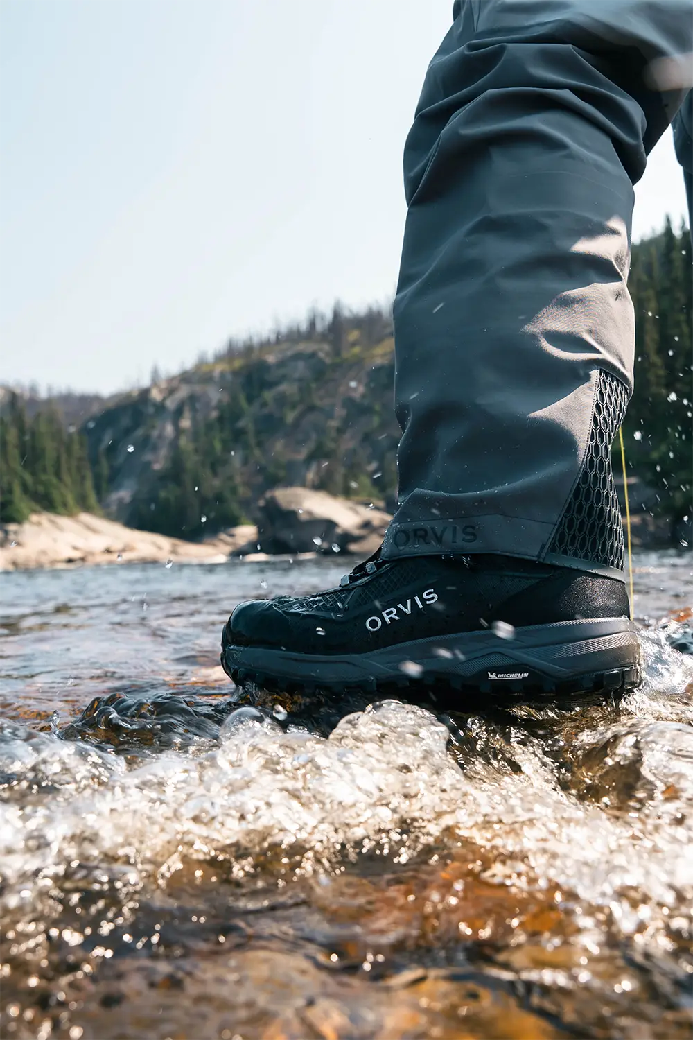 Close-up of a person wearing black Orvis boots and grey pants standing in a shallow rocky river with water splashing.