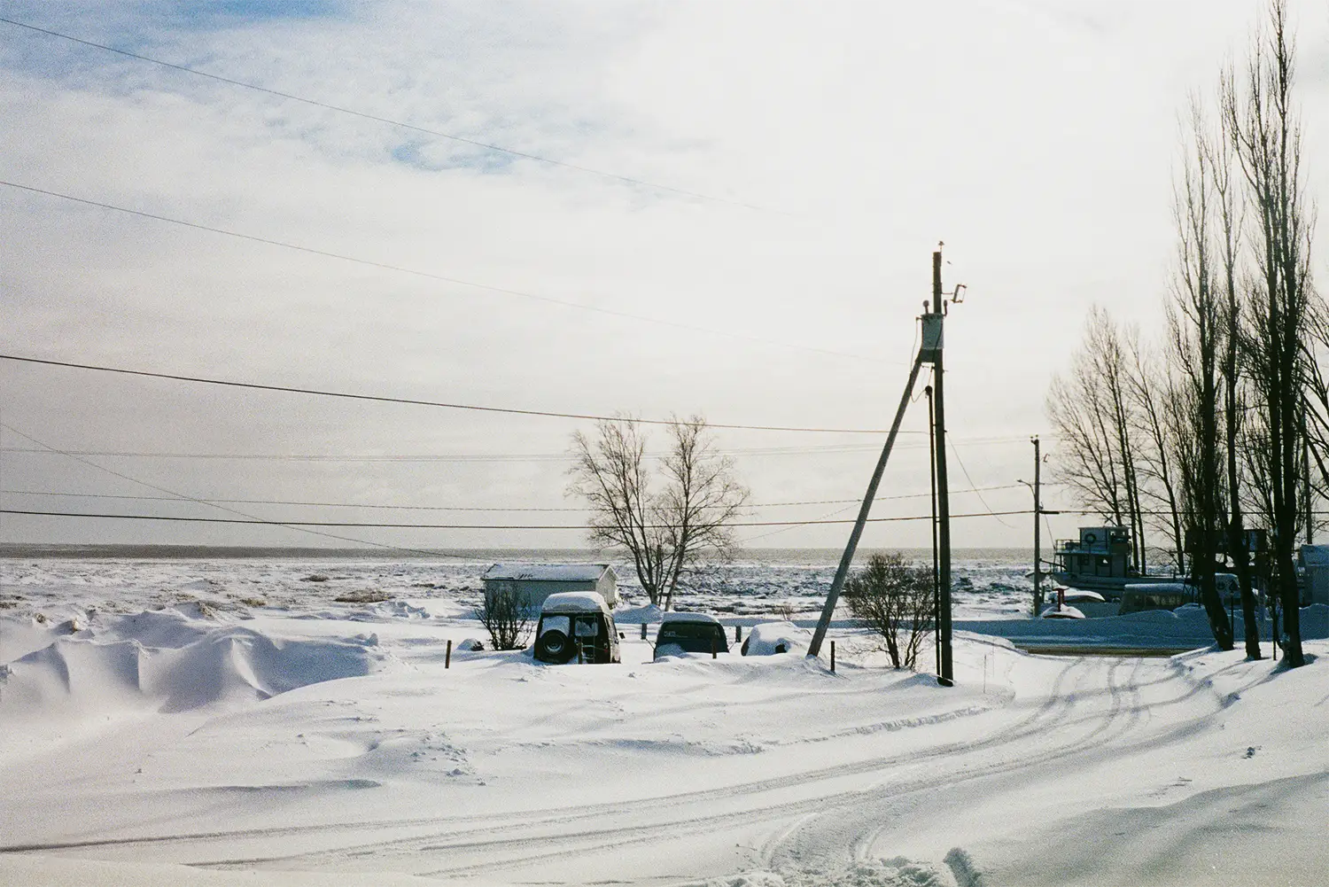 Snow-covered parked cars, leafless trees, and utility poles along a snow-covered road with a boat and frozen landscape in the background.