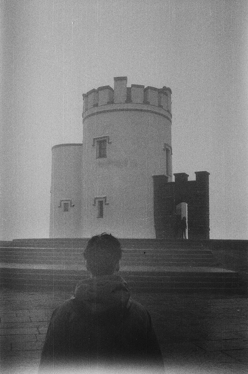 Grainy black-and-white photo of a person with their back to the camera facing a round tower with battlements and stairs leading up to it.