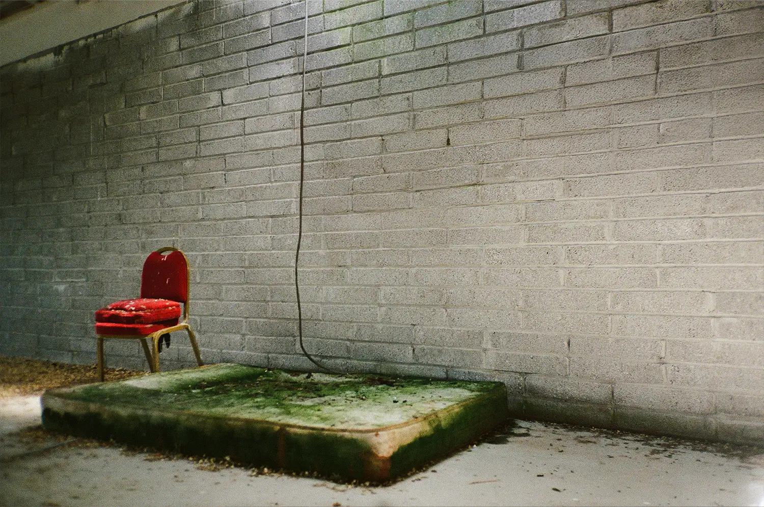 Red chair with cushion next to a moldy mattress on the floor against a gray brick wall.