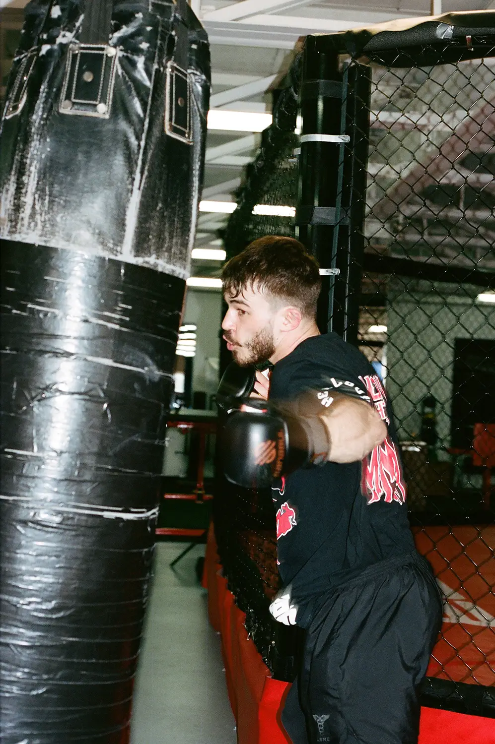 Man wearing black boxing gloves punching a black heavy bag inside a gym with chain-link fence.