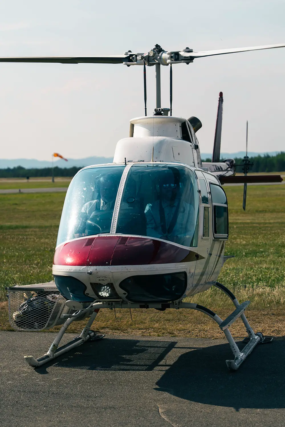 Front view of a small white and red helicopter with two pilots inside, parked on a tarmac with a grassy field in the background.