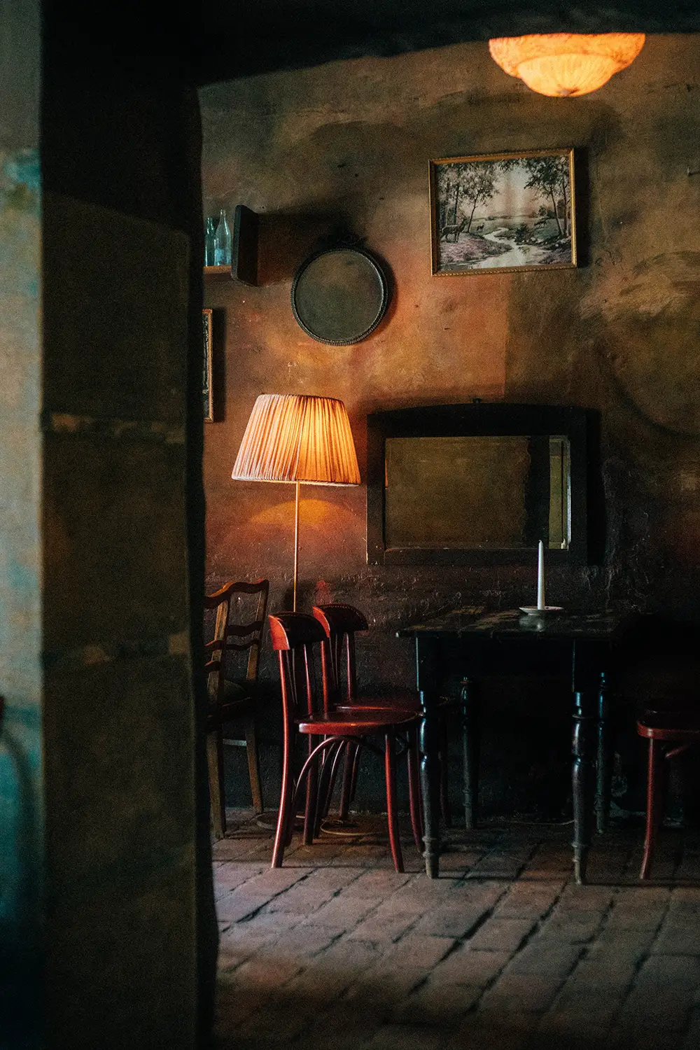Dimly lit rustic room with red wooden chairs, a candle on a dark wooden table, a standing lamp, and framed wall art.