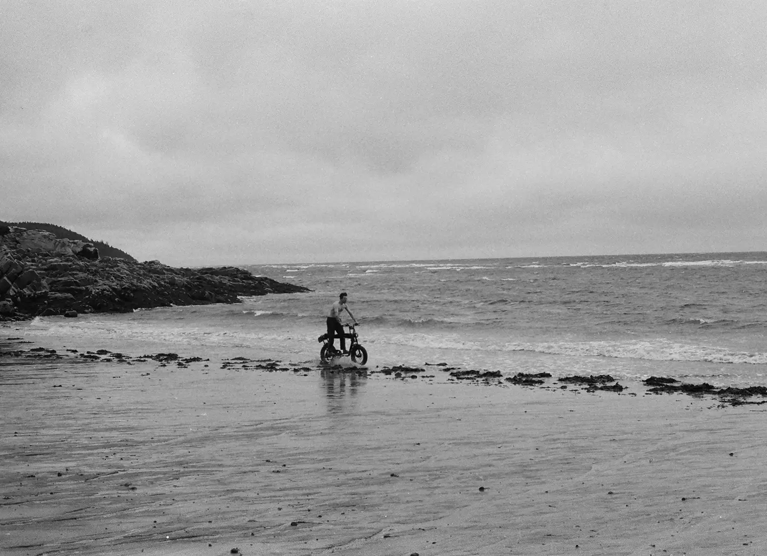 Man riding a bicycle near the shoreline on a cloudy day with rocky coast in the background.