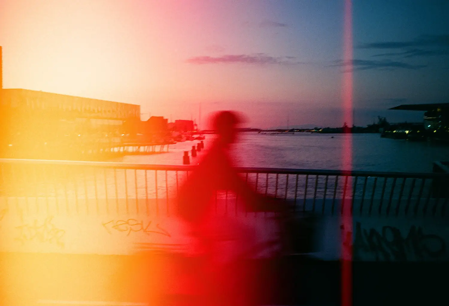 Blurred silhouette of a person riding a bicycle on a waterfront bridge during sunset with a colorful light leak effect.