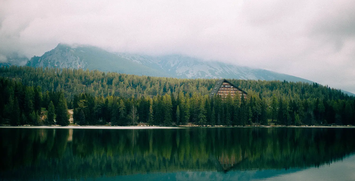A triangular lodge nestled among dense pine trees on the shore of a calm lake with misty mountains in the background.